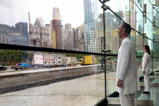 Photo documentation from Ernesto Pujol's project, 9-5 in 2015. Two performers in white clothing look out to a city landscape from inside a glass walled building.