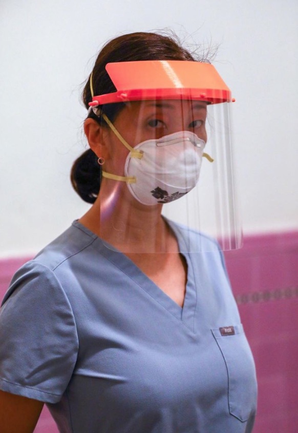A woman in blue scrubs wears a protective face shield and face mask