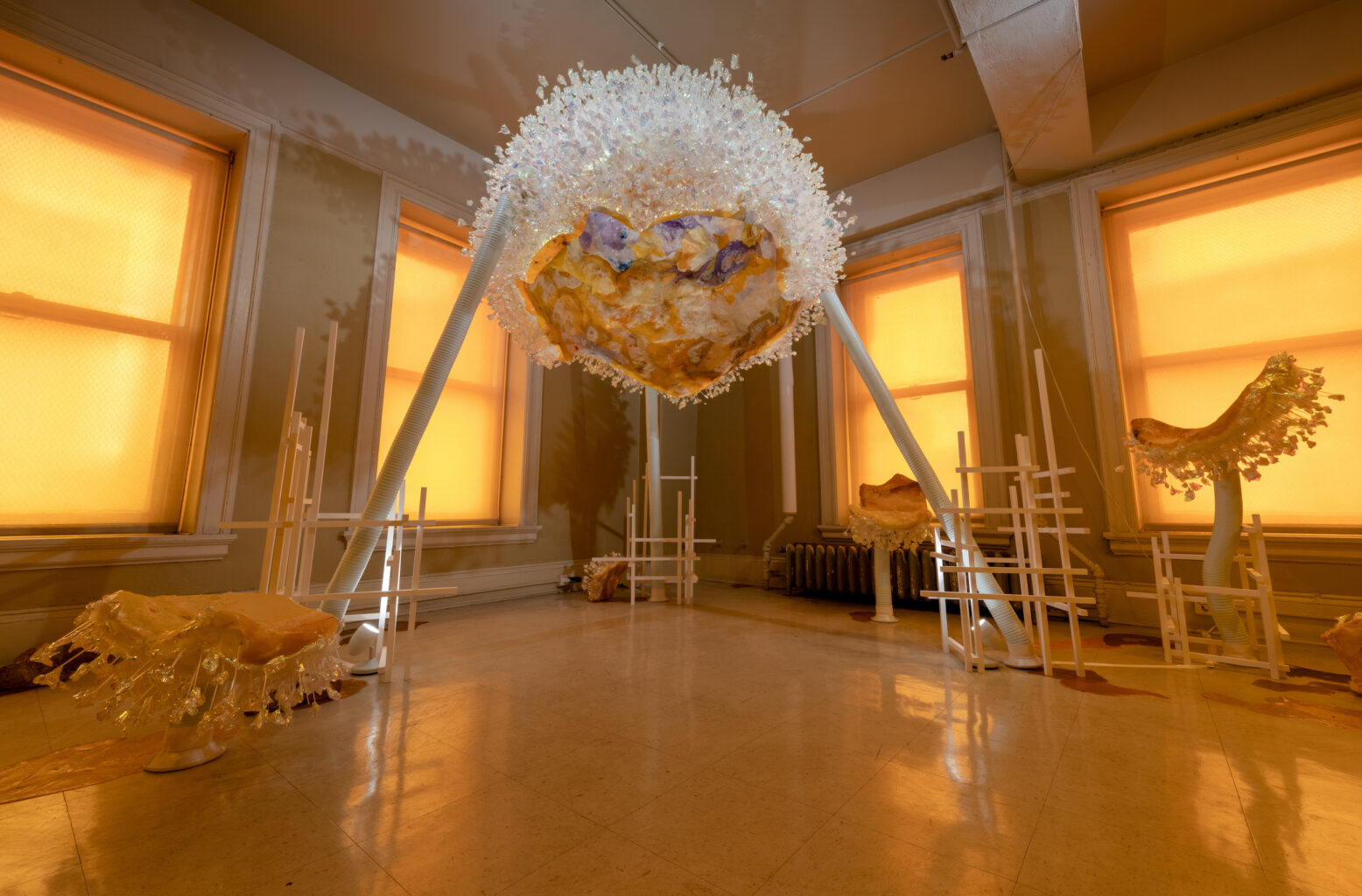 A amber-tinted room with four windows that seem to be glowing. A large sculpture looms in the corner in the room, like a white bedazzled alien on two legs with a round body. There are mushroom-like smaller sculptures that surround it, also bedazzled with shiny hanging objects.