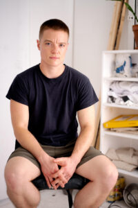 A portrait photo of Nolan Hanson. They are a caucasian trans person with a shaved head, wearing a black t-shirt and green shorts. They are sitting on a still in a workshop.
