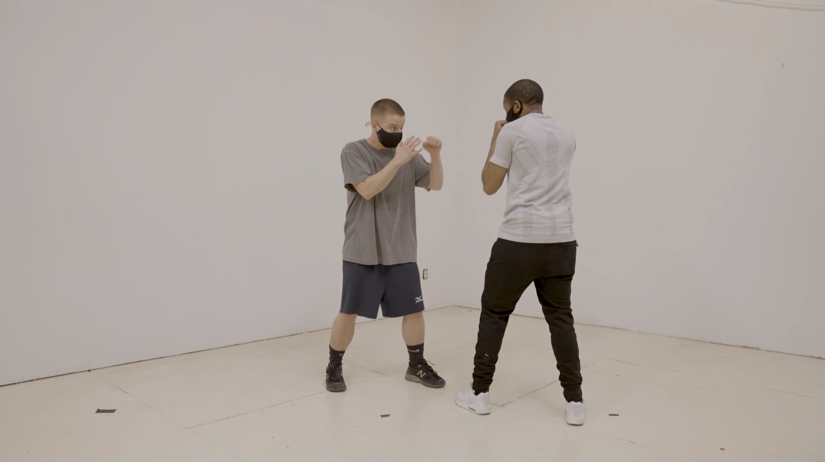 Two people stand in the corner of a white room, each in a boxing stance and wearing face masks.