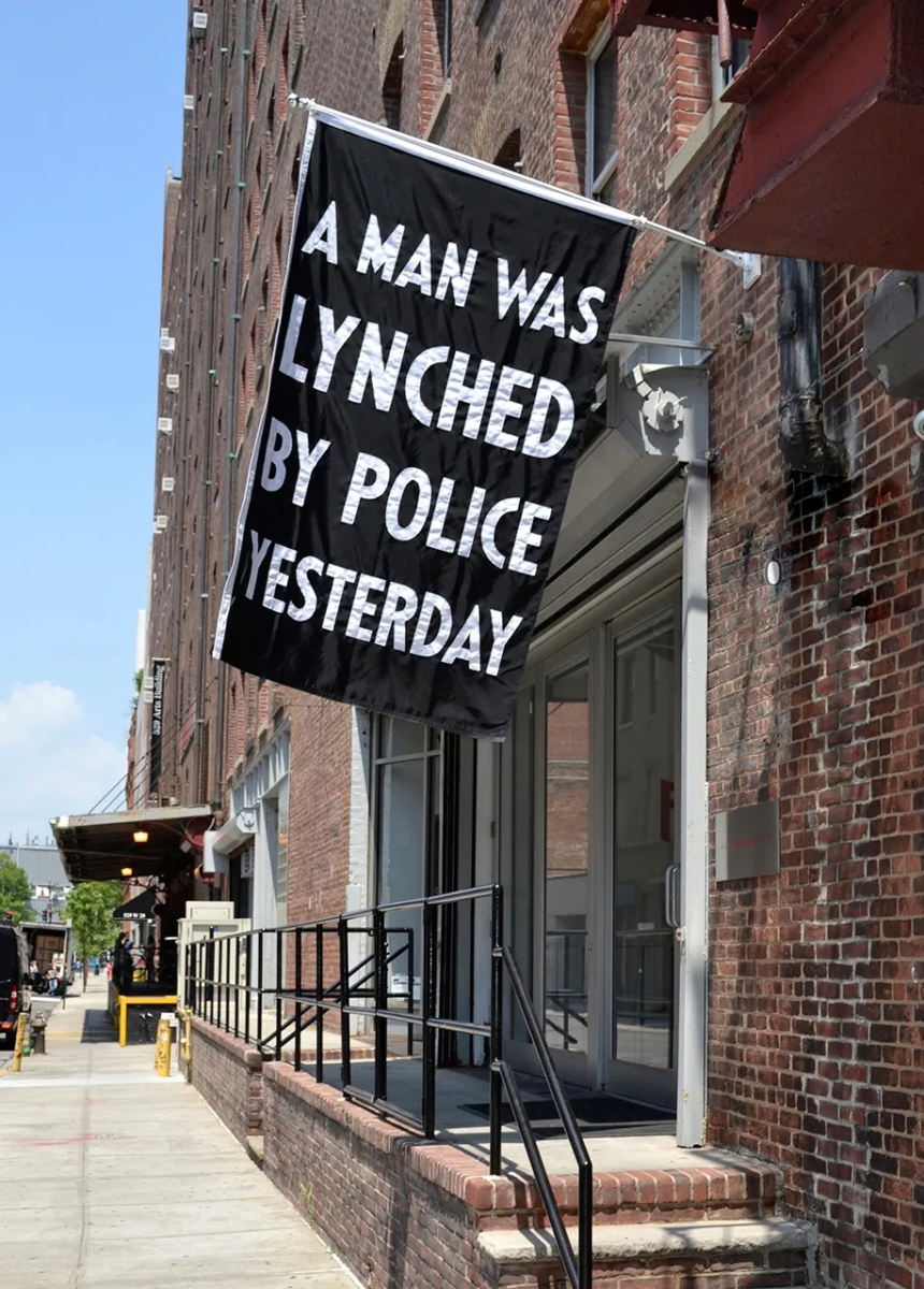 A brown brick city storefront building with a large banner flag hung at ceiling height outside. The banner reads "A man was lynched by police yesterday" in white capitialized letters on a black background.