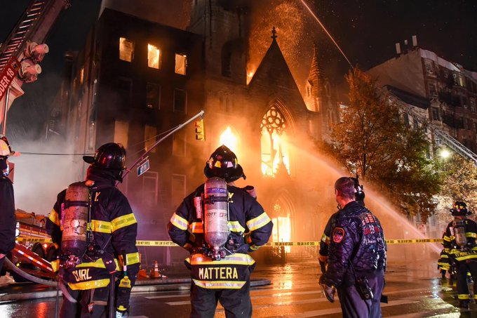 A photo of a church on fire with flames coming out of the front steeple and windows. There are three firemen in the foreground with their backs to the camera. A couple of fire hoses point their spray at the building