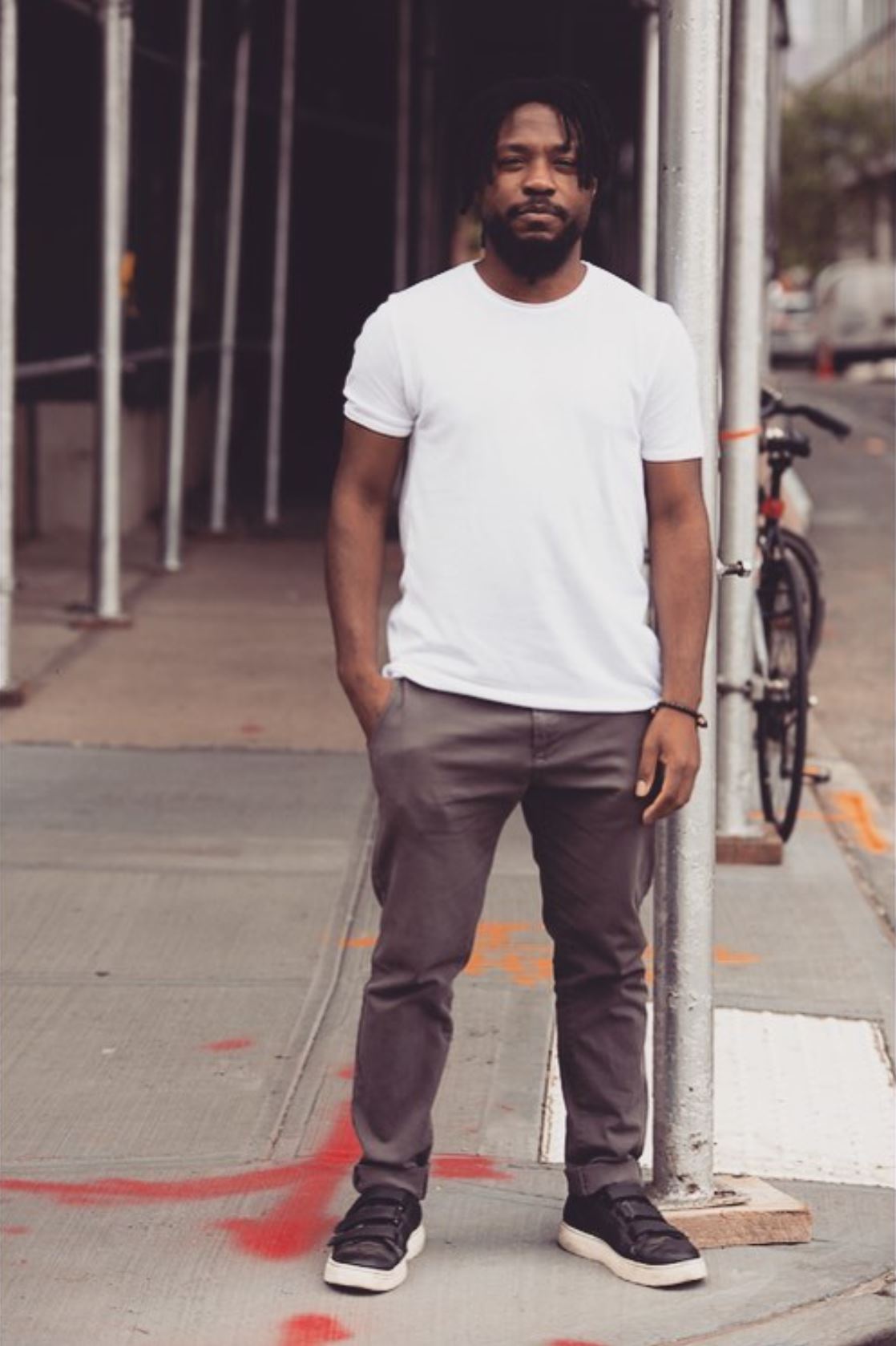 A Black man stands on the sidewalk of a city street. There is scaffolding poles behind him. He wears a white t-shirt and dark pants. He is looking at the camera, unsmilng, with a hand in his pocket. He is young with a short beard and short braided hair.