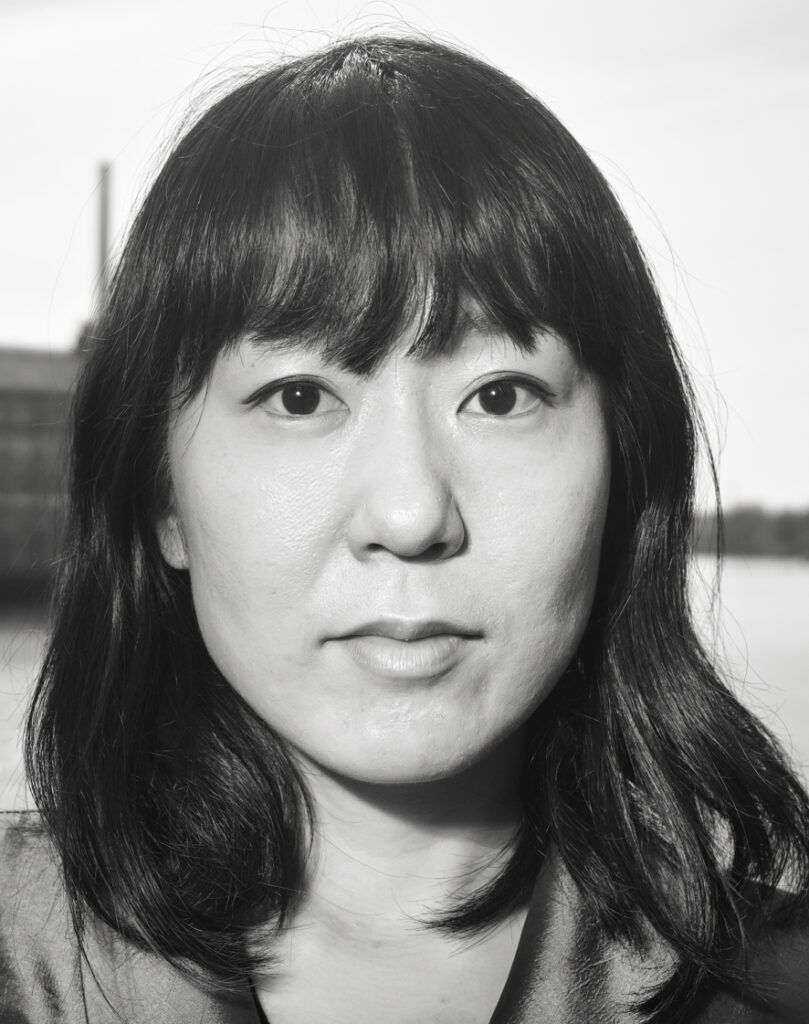 A black and white headshot of an Asian female presenting person with shoulder length dark hair and bangs.
