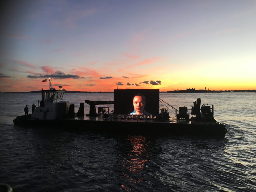 Screen on a boat during a sunset in the seashore. The screen shows the portrait of a person while the sky is full of colors.