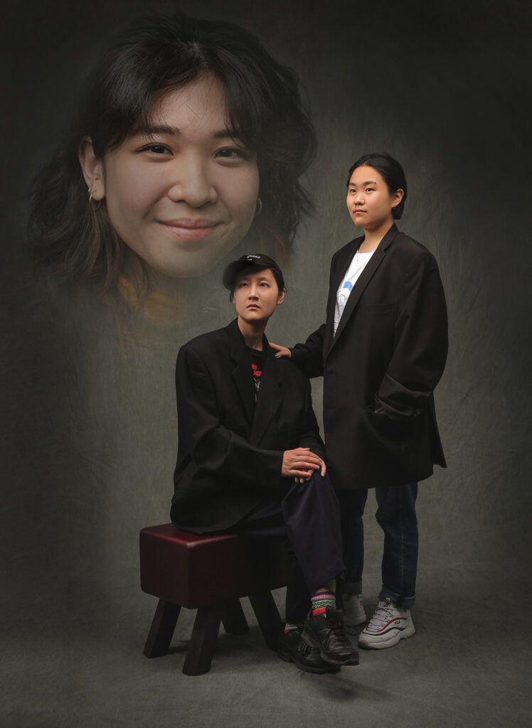 A photo of three people, staged like a traditional studio portrait with a grey photo backdrop. There are two people in the photo, a female presenting Asian person standing and a male presenting Asian person sitting on a stool, both wearing black. They are looking off frame into the distance. The third person is super imposed into the upper left quadrant of the photo with a vignette around their face. They are a female presenting Asian person with brown hair.