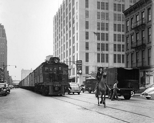 A historic photo of a city street with a street rail car and old cars on the street. Sharing the street is a man on a horse.