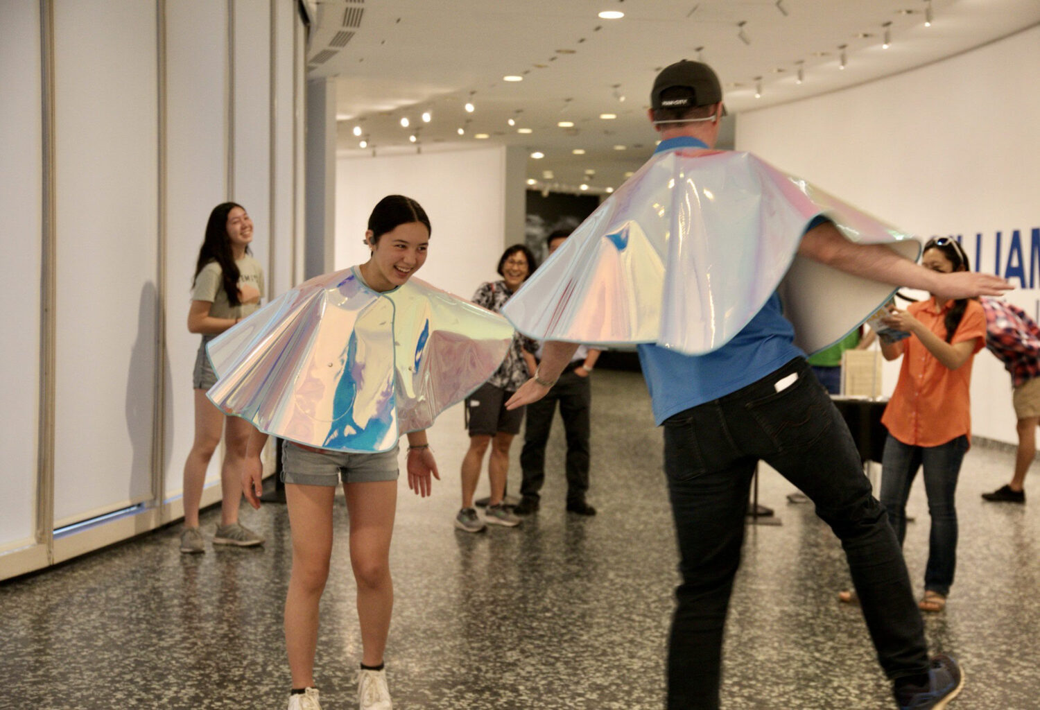 A photo inside what looks to be a circular hall with concrete floors and white walls. There are two people in the foreground dancing around eachother wearing iridescent capes that cover their arms down to their waists. There is a group of onlookers in the background smiling and taking photos of them.