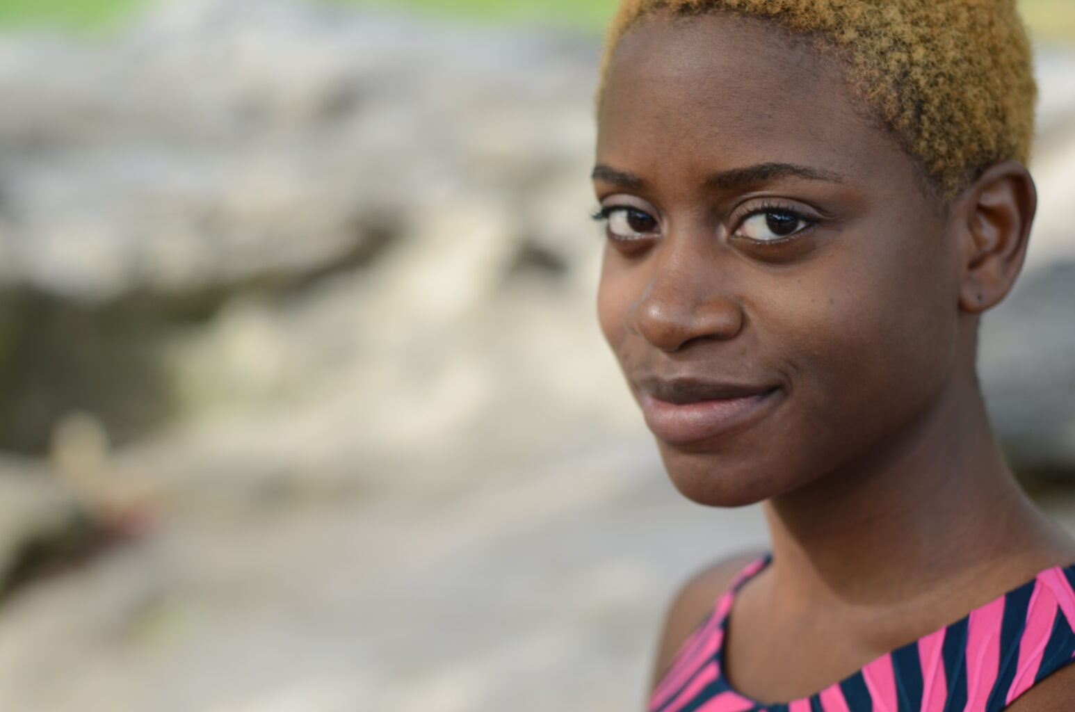 A headshot of Katrina Reid. She is a Black femme woman with short, almost shaved bleached hair, smiling while looking at the camera, her face pointed away to the left. There is a blurred landscape behind her that looks like a rock formation. She is wearing a pink and black shirt.